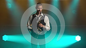 A young retro stylish guy plays on the golden shiny saxophone in the turquoise spotlights on stage. Dark studio with