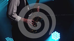 A young retro stylish guy plays on the golden shiny saxophone in the turquoise spotlights on stage. Dark studio with