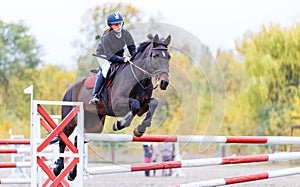 Young rider girl on bay horse jumping over barrier