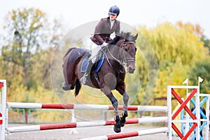 Young rider girl on bay horse jumping over barrier