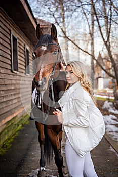 Young rider woman blonde with long hair in white clothes standing near brown horse