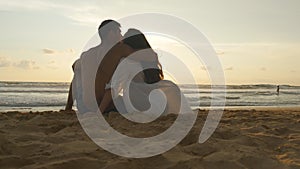 Young romantic couple is enjoying beautiful sunset sitting on the beach and hugging. A woman and a man sits together in