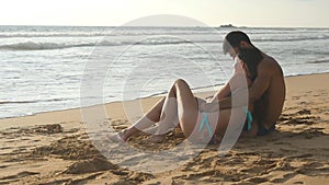 Young romantic couple is enjoying beautiful view sitting on the beach and hugging. A woman and a man sits together in