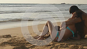 Young romantic couple is enjoying beautiful view sitting on the beach and hugging. A woman and a man sits together in