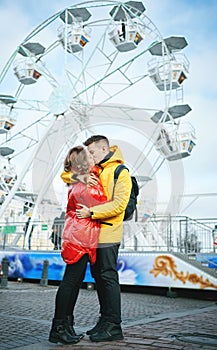 Young romantic couple kissing, hugging, walking on the street, wearning in bright down jackets. ferris wheel on