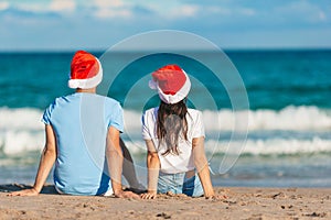 Young romantic couple in red Santa hats sitting on tropical white sand beach celebrating Christmas