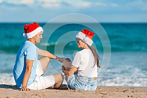 Young romantic couple in red Santa hats sitting on tropical white sand beach celebrating Christmas