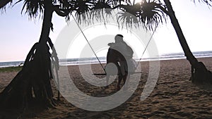 Young romantic couple sitting together on swing at the tropical sea beach and hugging. Pair in love relaxing and kissing