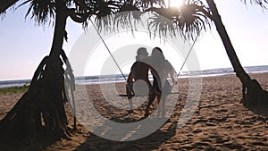 Young romantic couple sitting together on swing at the tropical sea beach and hugging. Pair in love relaxing and kissing