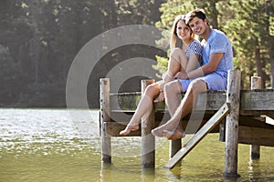 Young Romantic Couple Sitting On Wooden Jetty Looking Out Over L