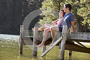 Young Romantic Couple Sitting On Wooden Jetty Looking Out Over Lake