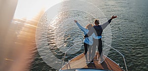 Young romantic couple standing on the prow of a yacht with hands up  during sunset. Sea travel