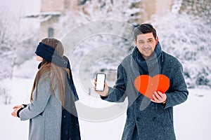 Young romantic couple, surprise outdoor, winter time. Man holding red heart and mobile phone with blank empty screen
