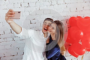 Young romantic loving beautiful couple guy with a girl with red heart shaped air balloons take a selfie on the phone on an