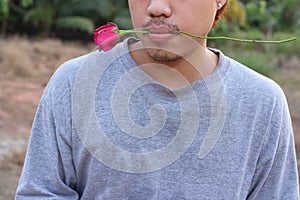 Young romantic man is holding a red rose in his mouth on nature blurred background. Love and romance Valentine`s day concept.