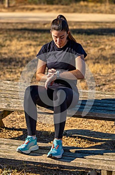 Young runner girl in sportswear sitting on a wooden bench in a park, enjoying listening to music with white wireless headphones