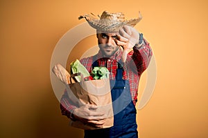 Young rural farmer man holding fresh groceries from marketplace over yellow background with angry face, negative sign showing