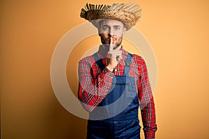 Young rural farmer man wearing bib overall and countryside hat over yellow background asking to be quiet with finger on lips