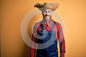 Young rural farmer man wearing bib overall and countryside hat over yellow background with a happy and cool smile on face