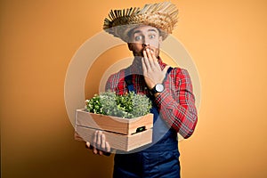 Young rural farmer man wearing countryside hat holding green organic plant from harvest cover mouth with hand shocked with shame