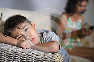 Young sad and bored Asian child at home couch feeling frustrated and unattended while mother networking on mobile phone as