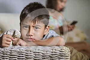 Young sad and bored Asian child at home couch feeling frustrated and unattended while mother networking on mobile phone as