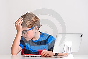 Young, sad boy sitting at desk at the table with his head in his hands. Stressed kid with hand on head. Education online