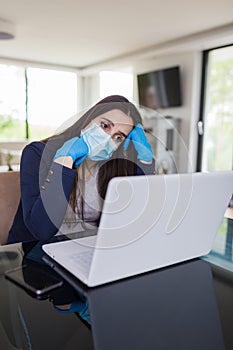 Young sad businesswoman working on laptop from home, wearing protective mask and gloves. Coronavirus quarantine and business