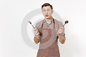 Young sad concerned man chef or waiter in striped brown apron, shirt holding fork, spoon isolated on white background