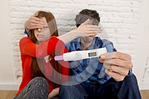 Young sad couple in shock and surprise holding pink positive pregnancy test blocking eyes scared