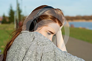 Young, sad cry redhead girl in the spring in the park near the river listens to music through wireless bluetooth headphones