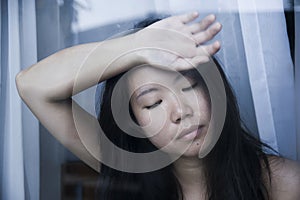 Young sad and depressed Asian Chinese woman looking thoughtful through window glass suffering pain and depression in sadness conce