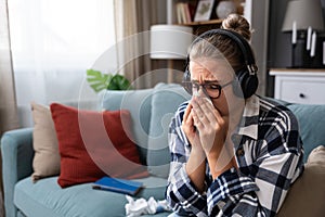 Young sad girl sitting alone at home on sofa listening to sad music on wireless headphones crying and wiping her nose with a paper