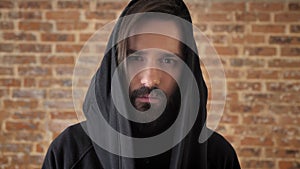 Young sad man with beard in hood is watching at camera, emotional concept, brick background