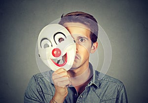 Young sad man hiding behind happy clown mask