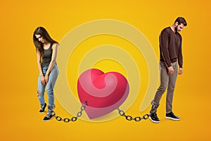 Young sad man and woman wearing casual clothes chained to pink heart on yellow background