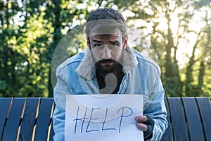 Young sad sick poor homeless man sitting on the bench on the street holding help sign paper feeling lonely and depressed because