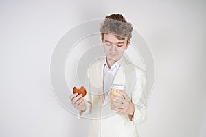 Young sad student guy in a business suit stands with a Cup of coffee and cookies in his hands on a white background in the Studio