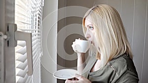 Young sad thinkful beautiful blond woman standing near the window with blinds in the morning and drinking coffee