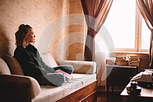 Young sad woman holding book looking through window while sitting on sofa in living room