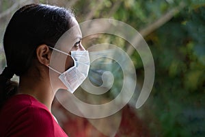 Young sad woman wearing a face mask looking through the window with her reflection on the glass. Coronavirus and Quarentine