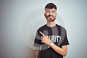 Young safeguard man with tattoo wering security uniform over isolated white background cheerful with a smile of face pointing with