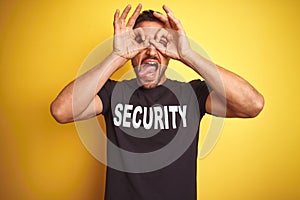 Young safeguard man wearing security uniform over yellow isolated background doing ok gesture like binoculars sticking tongue out,