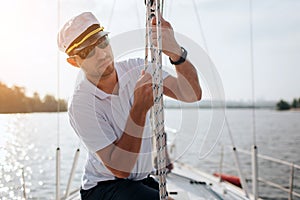 Young sailor in sunglasses and cap holds and moves ropes with both hands. He is calm and concentrated. Young man is