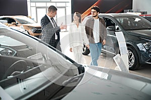 Young sales man talking with man and woman customers helps to choose automobile