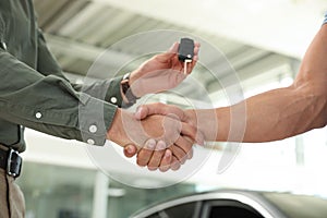 Young salesman shaking hands  customer and giving car key in modern auto dealership, closeup
