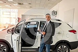 young salesman in suit standing near at car in new modern dealership