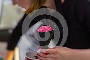 Young saleswoman in an ice cream parlor putting Bright pink fruit ice cream balls in Black waffle cone. Selective focus