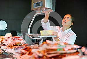 Young saleswoman weighing piece of semi-hard cheese on scales