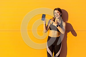 Young satisfied beautiful sporty woman in black sportwear standing near orange wall background and holding phone, pointing finger